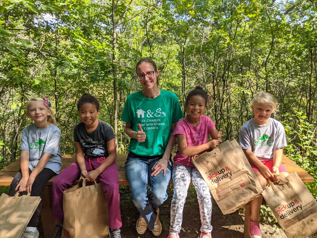 A parent volunteer in a St. Charles Home and School Council t-shirt sits outdoors with four smiling students holding grocery bags, celebrating a school volunteer event.